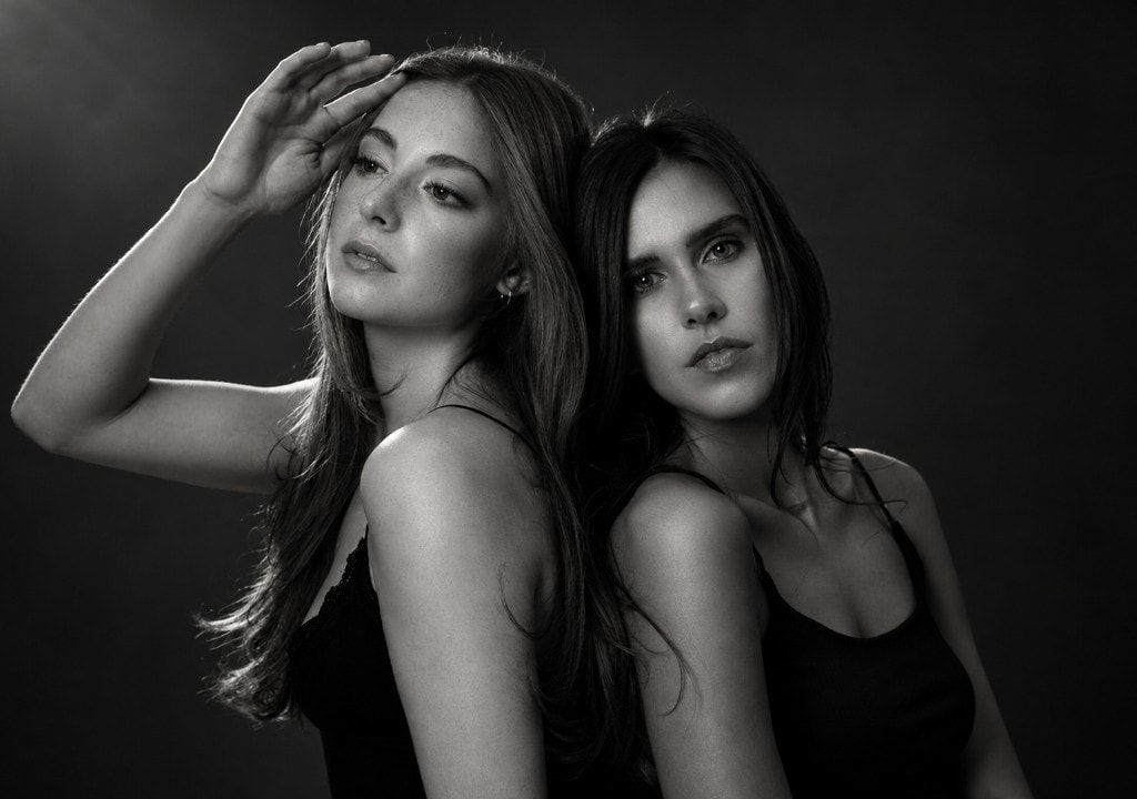 Black and white studio portrait of two women standing back to back, one with a hand raised to her forehead