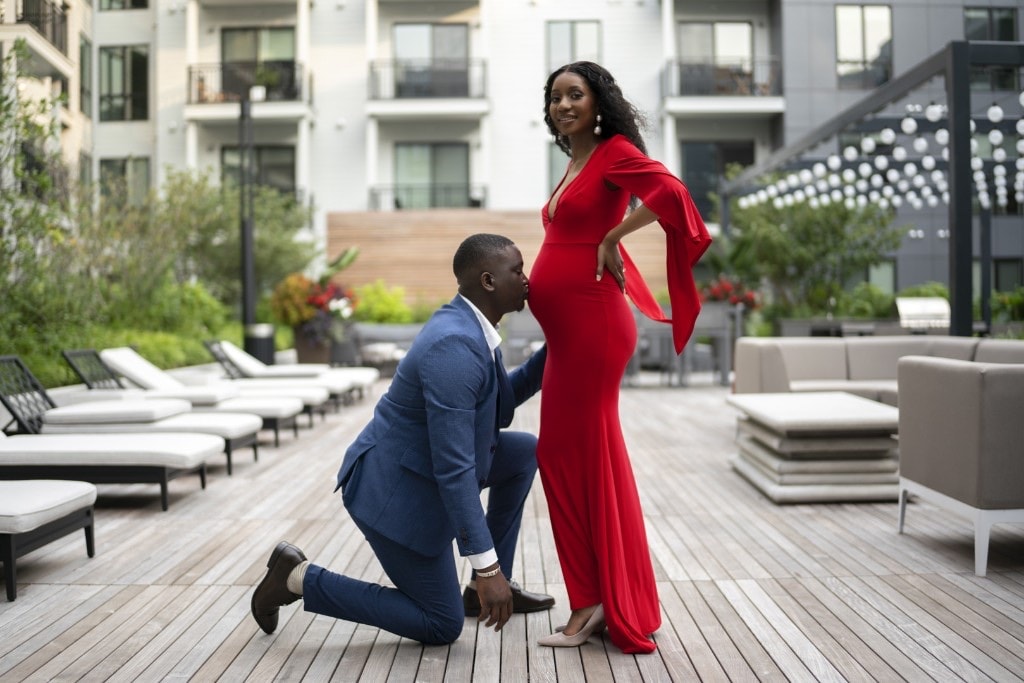 Man in a blue suit kneeling to kiss the belly of his pregnant partner in a red gown on a rooftop