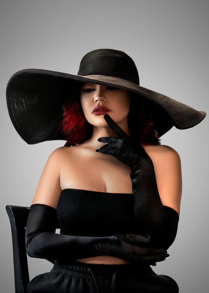 Woman with red hair in a wide black hat and long black gloves, one finger raised to her lips